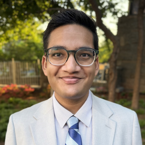 PhD Student Riyadh Baksh Smiles at the Camera against a group of trees outside. 